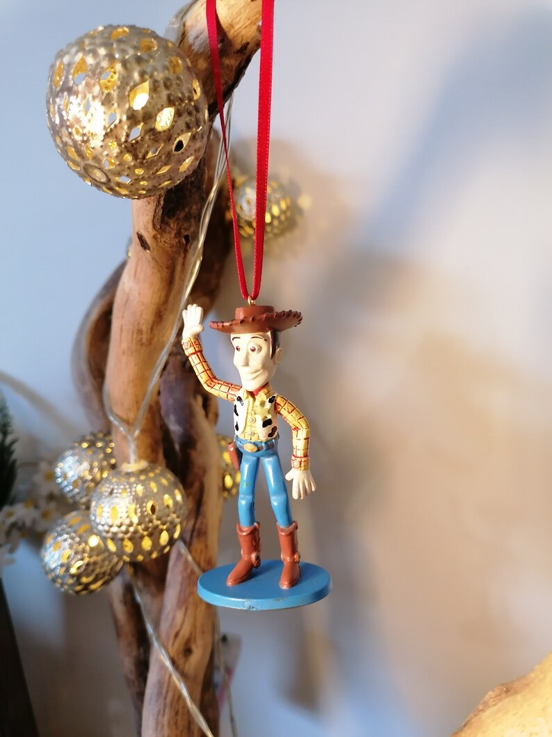 May include: A red string hangs from a wooden branch, holding a plastic figurine of Woody from Toy Story. Woody is wearing his signature cowboy hat, vest, and jeans. He is waving with his right hand and has a friendly expression on his face.