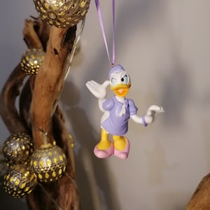 May include: A purple and white plastic figurine of a cartoon duck wearing a purple dress and a white hat. The duck is holding out its right wing and has a friendly expression on its face. The figurine is hanging from a purple ribbon.