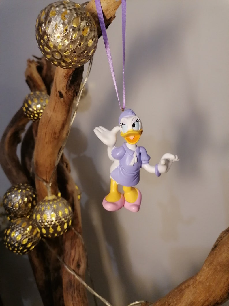 May include: A purple and white plastic figurine of a cartoon duck wearing a purple dress and a white hat. The duck is holding out its right wing and has a friendly expression on its face. The figurine is hanging from a purple ribbon.