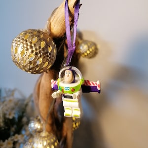 May include: A purple and white Christmas bauble featuring a Lego figurine of Buzz Lightyear from the Toy Story film franchise. The bauble is hanging from a purple ribbon.