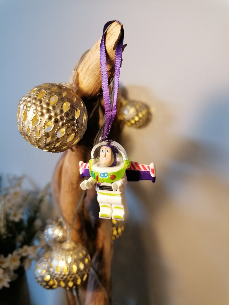 May include: A purple and white Christmas bauble featuring a Lego figurine of Buzz Lightyear from the Toy Story film franchise. The bauble is hanging from a purple ribbon.