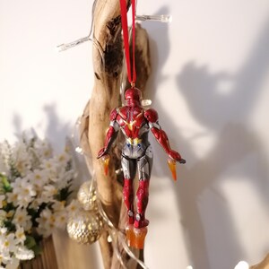 May include: Red and gold Iron Man Christmas ornament hanging from a red string. The ornament is a detailed figurine of Iron Man in flight.