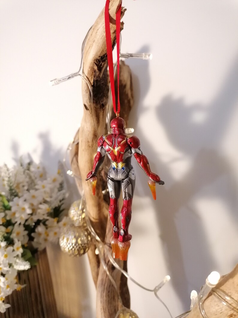 May include: Red and gold Iron Man Christmas ornament hanging from a red string. The ornament is a detailed figurine of Iron Man in flight.