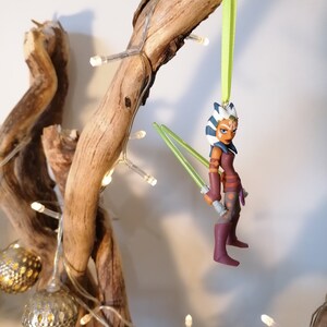 May include: A green, white, and blue Star Wars character ornament with green lightsabers hanging from a branch with white lights.