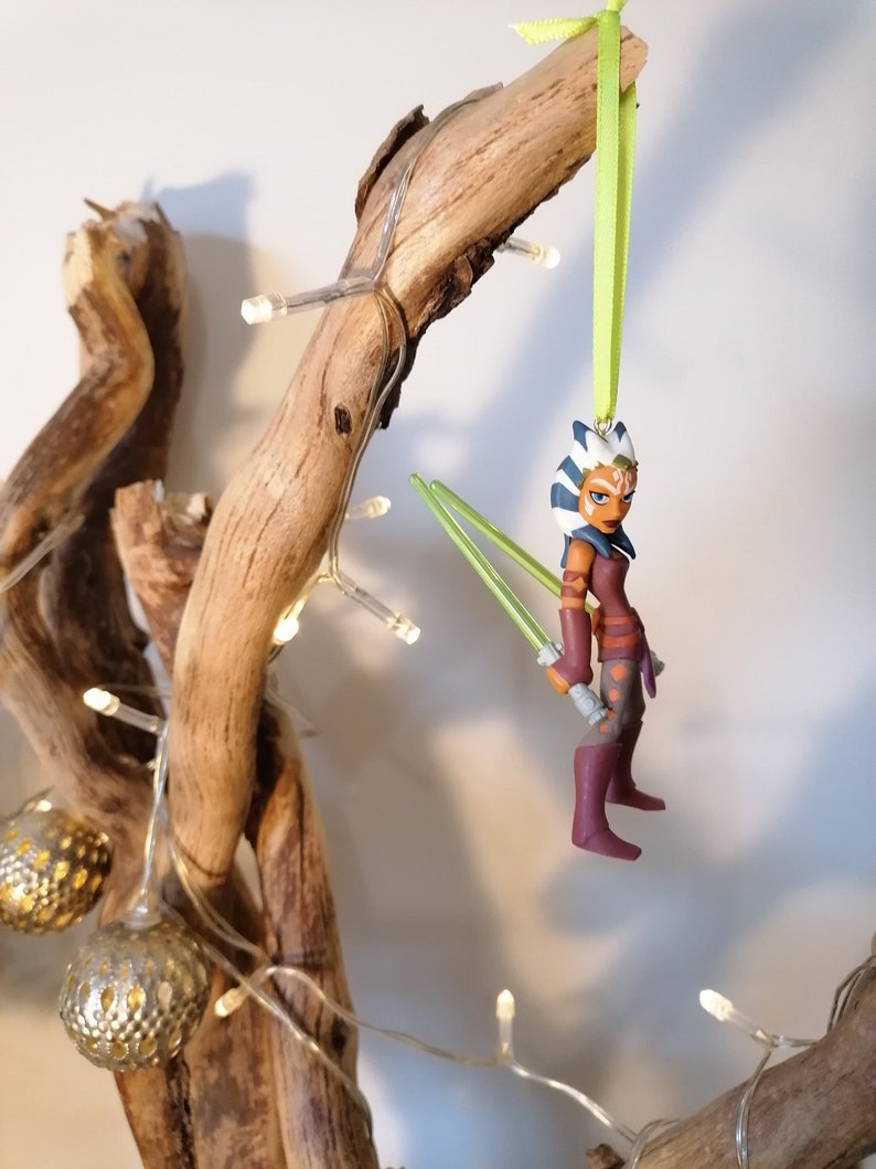 May include: A green, white, and blue Star Wars character ornament with green lightsabers hanging from a branch with white lights.
