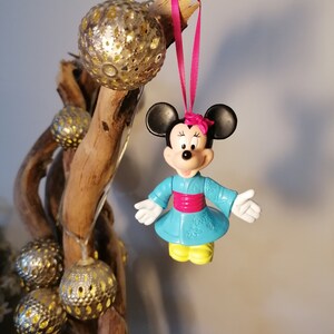 May include: A blue and pink Minnie Mouse ornament wearing a traditional Japanese kimono with a pink sash. The ornament is hanging from a pink ribbon on a branch with gold ornaments.