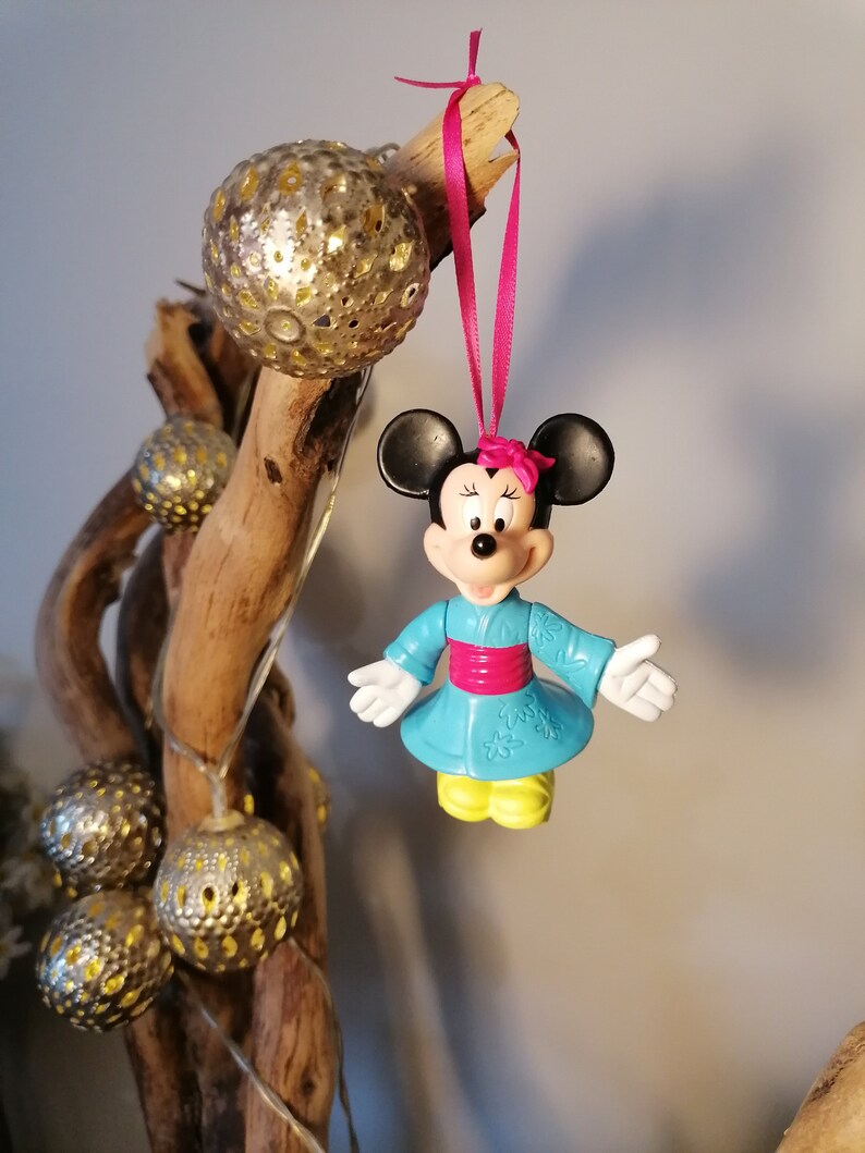 May include: A blue and pink Minnie Mouse ornament wearing a traditional Japanese kimono with a pink sash. The ornament is hanging from a pink ribbon on a branch with gold ornaments.