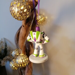 May include: A white, green, and purple Buzz Lightyear Christmas ornament. The ornament is hanging from a purple ribbon on a branch with gold and white lights.
