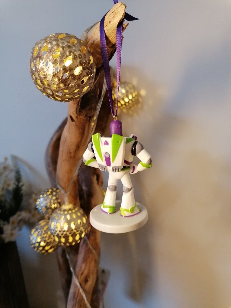 May include: A white, green, and purple Buzz Lightyear Christmas ornament. The ornament is hanging from a purple ribbon on a branch with gold and white lights.