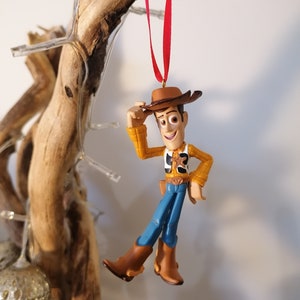 May include: A red string hangs from a wooden branch with a plastic toy ornament of Woody from Toy Story. Woody is wearing his signature cowboy hat, vest, and jeans. He is holding his hat in his right hand and smiling.