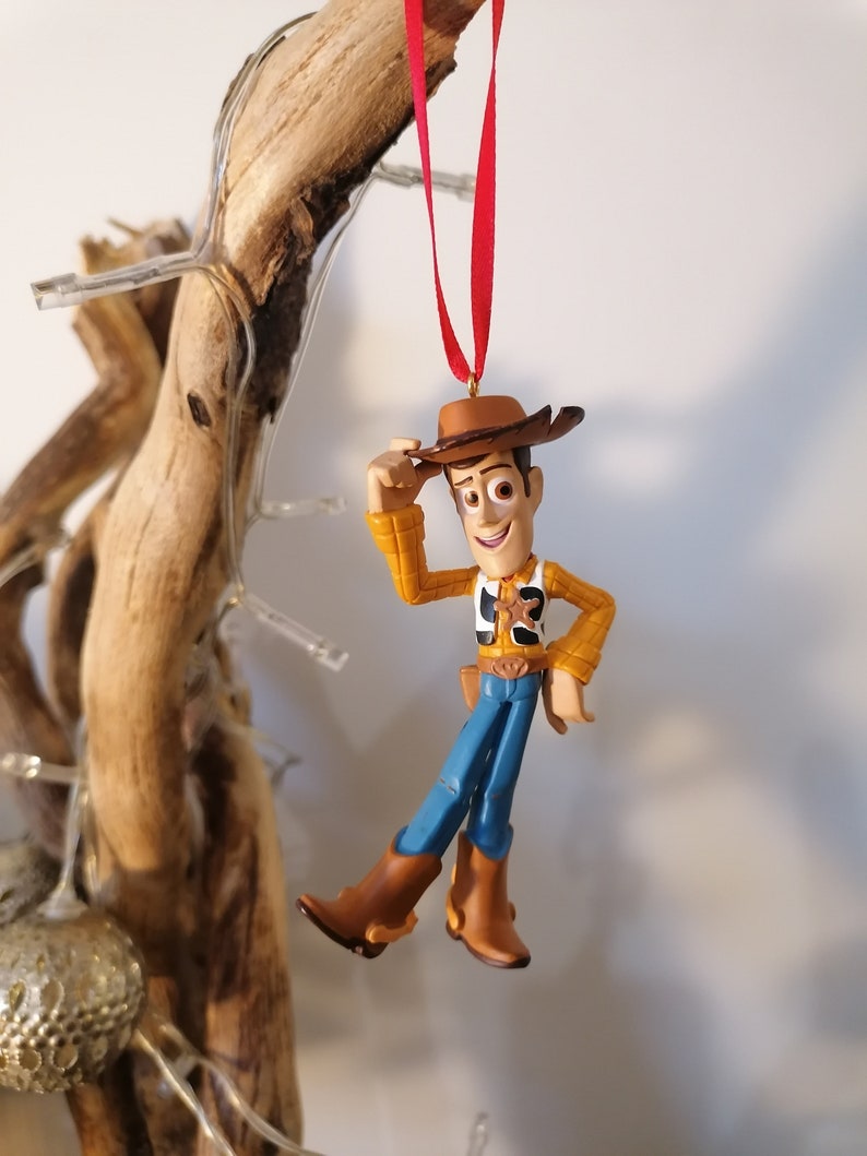 May include: A red string hangs from a wooden branch with a plastic toy ornament of Woody from Toy Story. Woody is wearing his signature cowboy hat, vest, and jeans. He is holding his hat in his right hand and smiling.