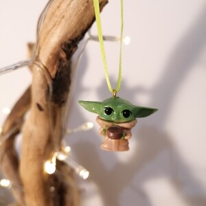 May include: A green and brown Christmas ornament featuring a character from the Star Wars franchise, The Mandalorian. The ornament is hanging from a green ribbon.