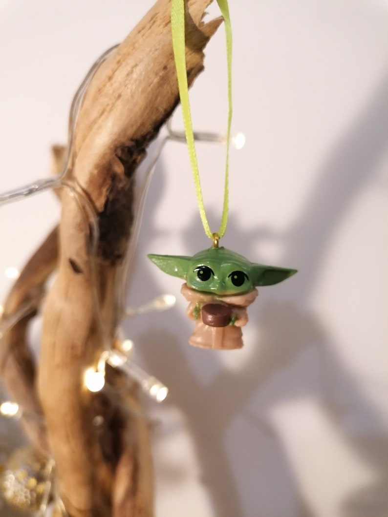 May include: A green and brown Christmas ornament featuring a character from the Star Wars franchise, The Mandalorian. The ornament is hanging from a green ribbon.