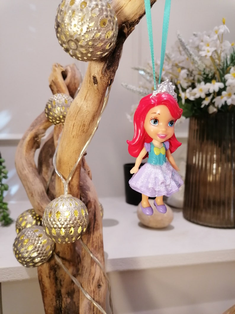 Peut inclure: Une petite poup&eacute;e en plastique aux cheveux roux, une robe violette et une couronne suspendue &agrave; une branche avec une guirlande lumineuse. La poup&eacute;e porte un haut bleu et vert et des chaussures violettes.