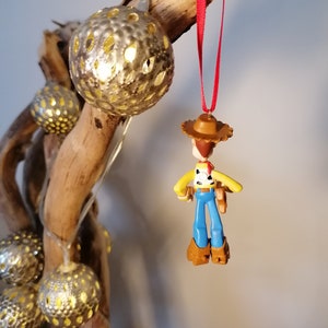 May include: A red string ornament featuring a plastic figurine of Woody from the Toy Story franchise. Woody is wearing his signature cowboy hat, vest, and jeans. The ornament is hanging from a branch with gold-colored decorative balls.