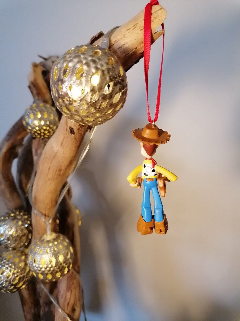 May include: A red string ornament featuring a plastic figurine of Woody from the Toy Story franchise. Woody is wearing his signature cowboy hat, vest, and jeans. The ornament is hanging from a branch with gold-colored decorative balls.