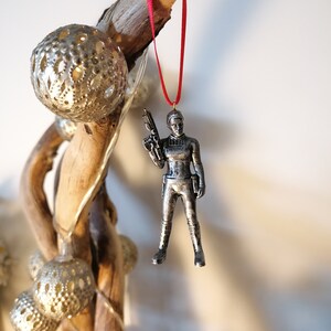 May include: A silver-coloured figurine of a woman holding a blaster, hanging from a red ribbon. The figurine is a Christmas ornament.
