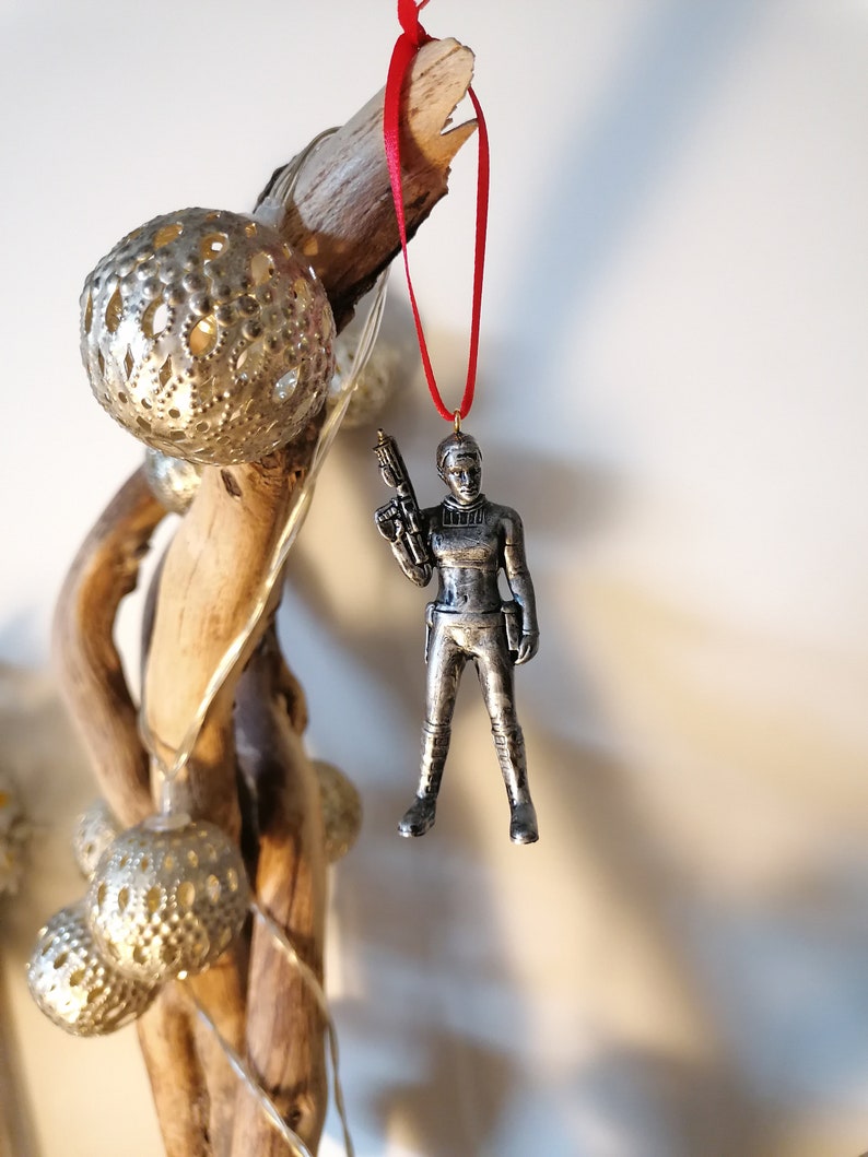 May include: A silver-coloured figurine of a woman holding a blaster, hanging from a red ribbon. The figurine is a Christmas ornament.