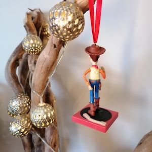 Puede incluir: Un adorno navide&ntilde;o rojo y blanco con una figura de pl&aacute;stico de Woody de Toy Story. Woody est&aacute; de pie en una plataforma roja con un disco negro. El adorno est&aacute; colgado de una cinta roja.