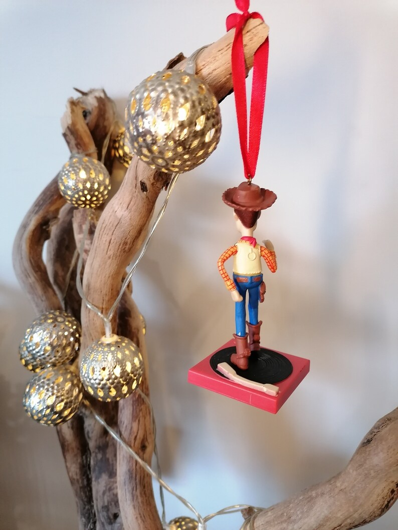 Puede incluir: Un adorno navide&ntilde;o rojo y blanco con una figura de pl&aacute;stico de Woody de Toy Story. Woody est&aacute; de pie en una plataforma roja con un disco negro. El adorno est&aacute; colgado de una cinta roja.