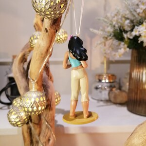 May include: A small figurine of a woman with long black hair wearing a blue and white outfit. She is standing on a round base and is hanging from a white string.