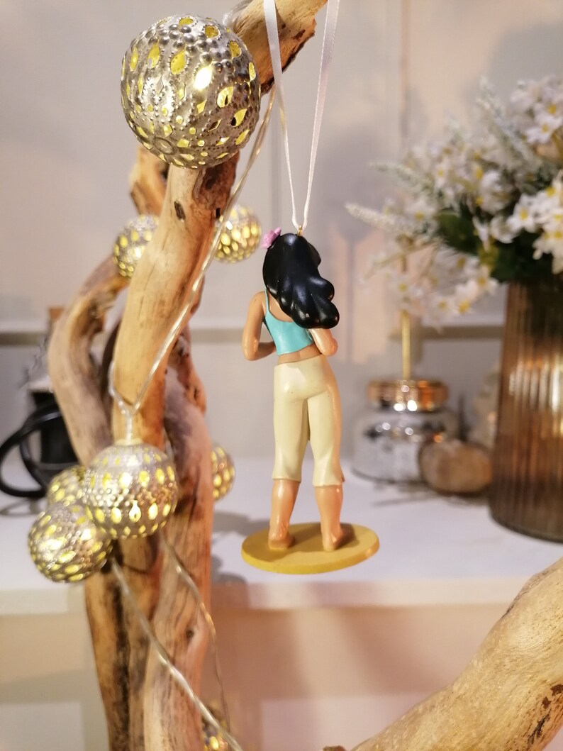 May include: A small figurine of a woman with long black hair wearing a blue and white outfit. She is standing on a round base and is hanging from a white string.