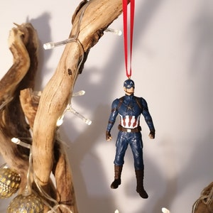May include: A red string hangs from a branch with a Captain America figurine ornament attached. The figurine is wearing a blue and red suit with a white star on the chest.