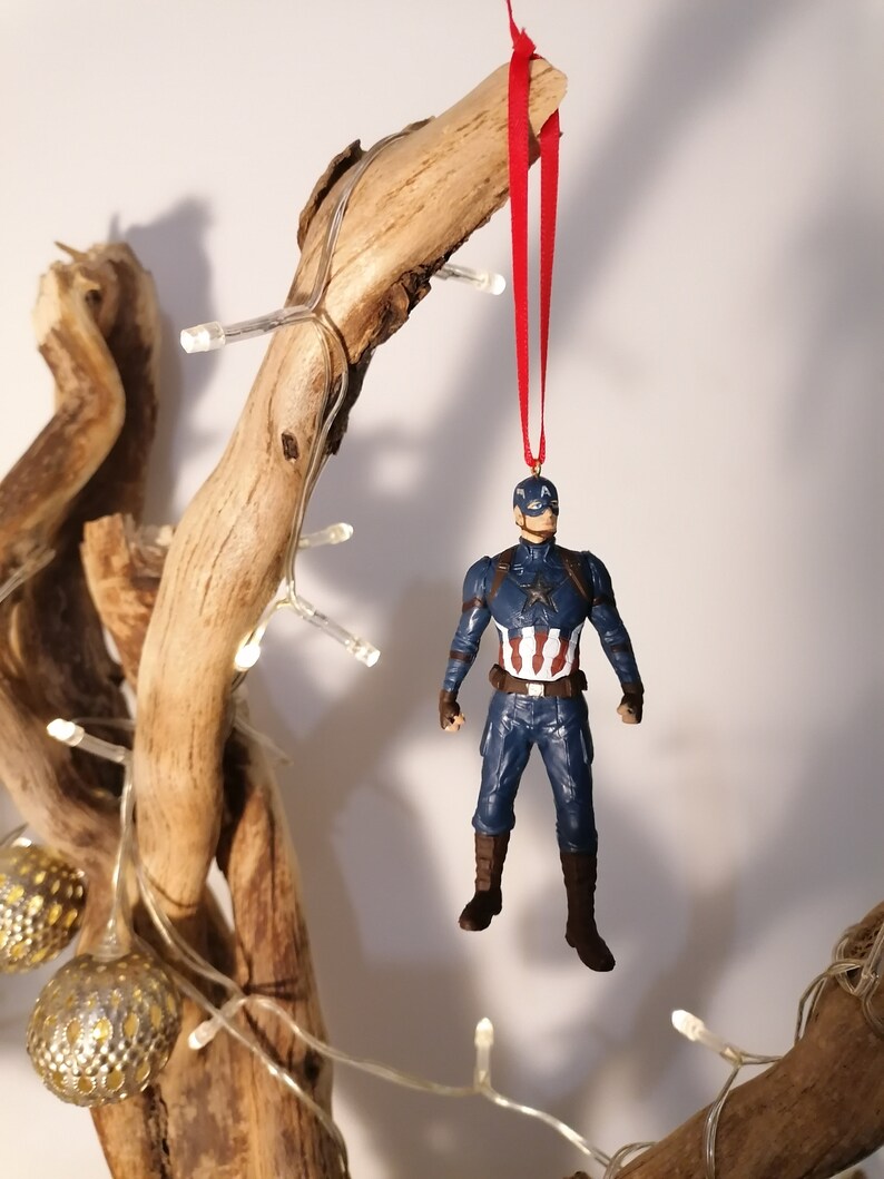 May include: A red string hangs from a branch with a Captain America figurine ornament attached. The figurine is wearing a blue and red suit with a white star on the chest.