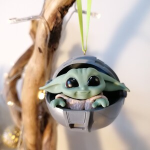 May include: A green and gray Christmas ornament featuring a small, smiling character in a round pod. The ornament is hanging from a green ribbon.