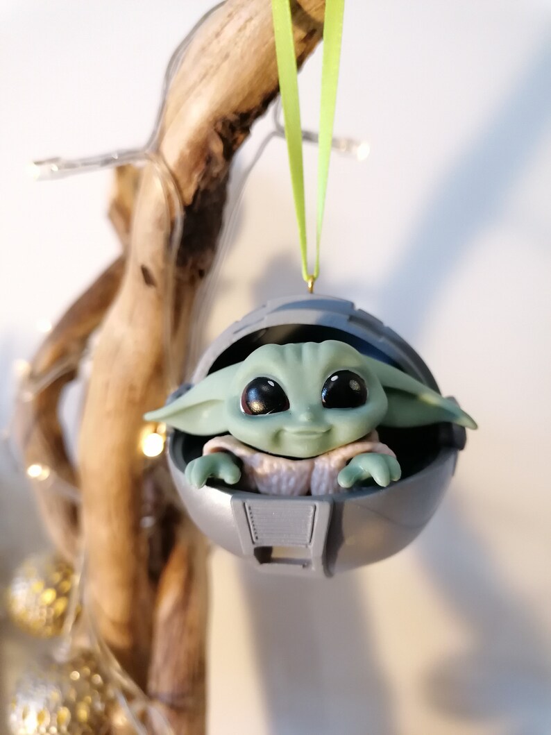 May include: A green and gray Christmas ornament featuring a small, smiling character in a round pod. The ornament is hanging from a green ribbon.