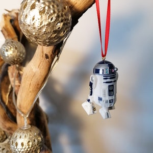 K&ouml;nnte beinhalten: Ein wei&szlig;er und blauer R2-D2 Star Wars Charakter Anh&auml;nger, der an einem roten Band an einem Zweig mit goldenen Dekorationskugeln h&auml;ngt.