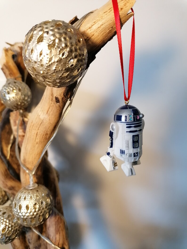 K&ouml;nnte beinhalten: Ein wei&szlig;er und blauer R2-D2 Star Wars Charakter Anh&auml;nger, der an einem roten Band an einem Zweig mit goldenen Dekorationskugeln h&auml;ngt.