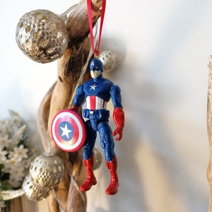 May include: A red, white, and blue Captain America action figure ornament with a red ribbon hanging from a branch. The figure is holding a red, white, and blue shield.