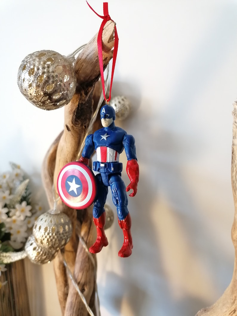 May include: A red, white, and blue Captain America action figure ornament with a red ribbon hanging from a branch. The figure is holding a red, white, and blue shield.