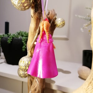 May include: A pink plastic doll ornament with orange hair hanging from a purple ribbon. The doll is wearing a pink dress and has a red crown on its head.