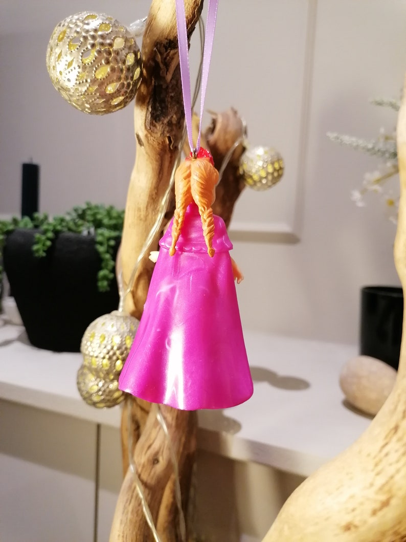 May include: A pink plastic doll ornament with orange hair hanging from a purple ribbon. The doll is wearing a pink dress and has a red crown on its head.