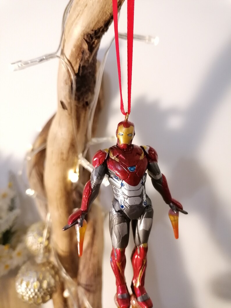 May include: Red and gold Iron Man Christmas ornament with a red ribbon. The ornament is a detailed figurine of Iron Man in flight.