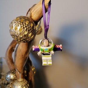 Puede incluir: Un adorno navideño de plástico morado y blanco con una figura Lego de Buzz Lightyear de la franquicia de películas Toy Story. El adorno está colgado de una cinta morada en una rama con luces decorativas doradas y plateadas.