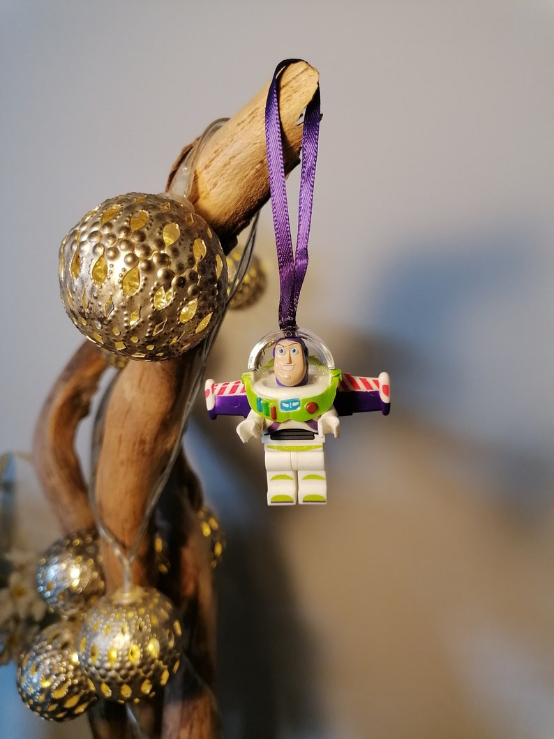 May include: A purple and white Christmas bauble featuring a Lego figurine of Buzz Lightyear from the Toy Story film franchise. The bauble is hanging from a purple ribbon on a branch with gold and silver decorative lights.