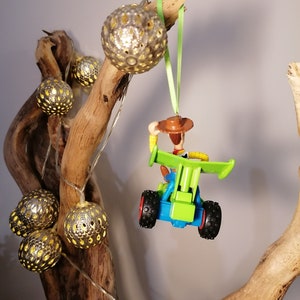 May include: A green and blue toy car with a cowboy figure driving it. The car is hanging from a branch with a green ribbon. The branch is decorated with silver and gold ornaments.