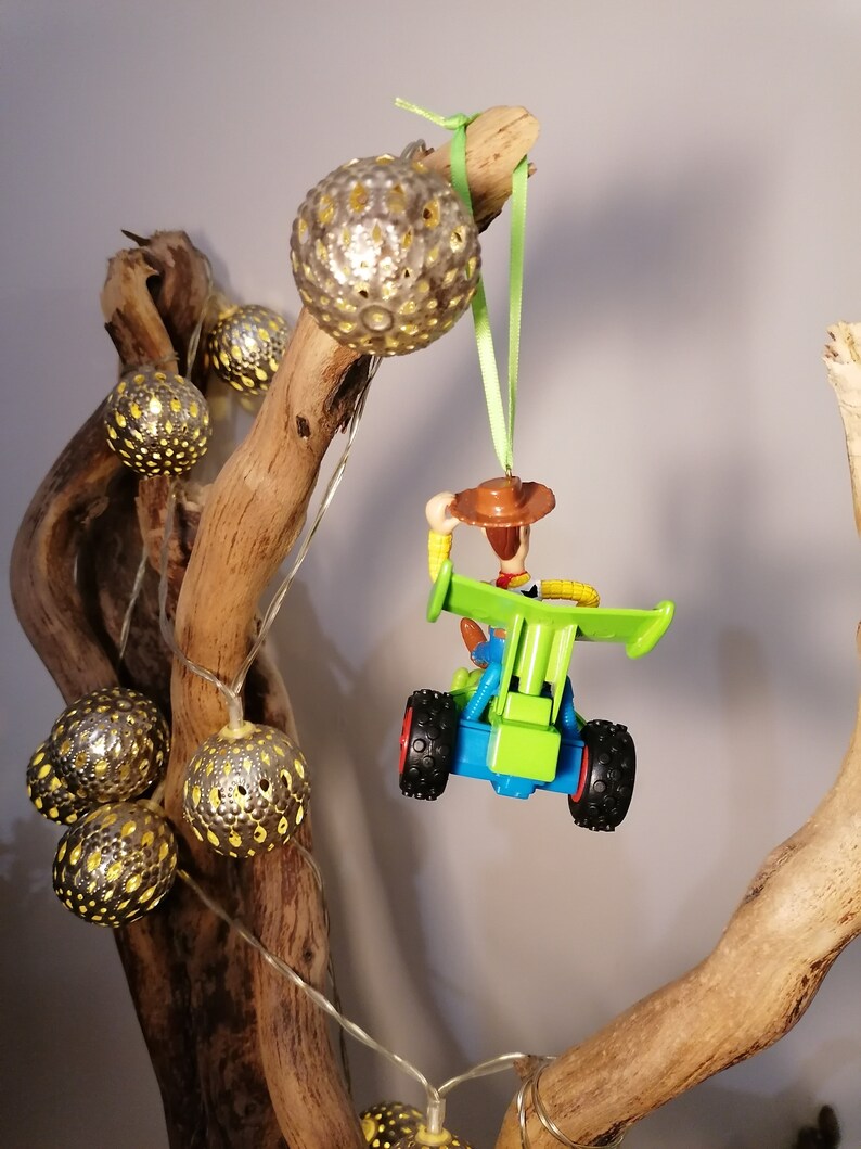 May include: A green and blue toy car with a cowboy figure driving it. The car is hanging from a branch with a green ribbon. The branch is decorated with silver and gold ornaments.