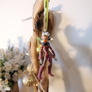 May include: A green and white Star Wars character ornament with a green lightsaber. The character is wearing a brown and purple outfit and has blue eyes. The ornament is hanging from a green ribbon.