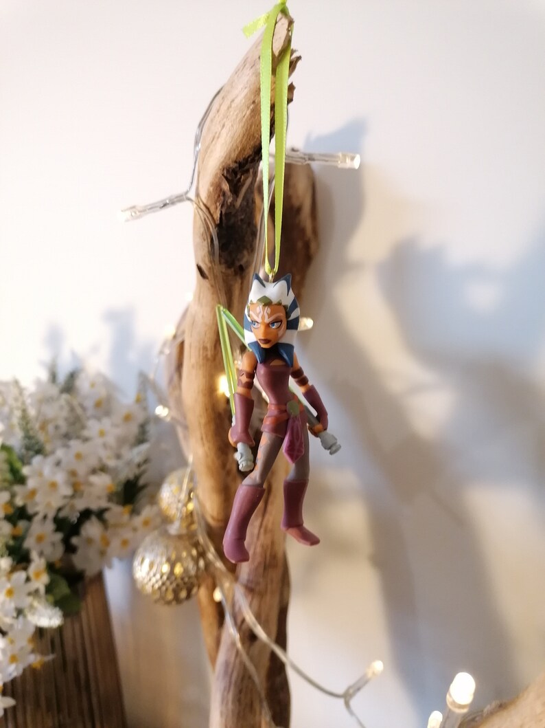 May include: A green and white Star Wars character ornament with a green lightsaber. The character is wearing a brown and purple outfit and has blue eyes. The ornament is hanging from a green ribbon.