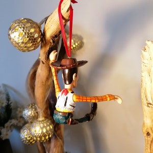 May include: A red string ornament featuring a plastic figurine of Woody from Toy Story. Woody is wearing his signature cowboy hat, vest, and jeans. He is posed with his arms outstretched and legs bent, as if he is climbing.