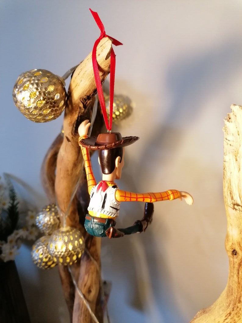 May include: A red string ornament featuring a plastic figurine of Woody from Toy Story. Woody is wearing his signature cowboy hat, vest, and jeans. He is posed with his arms outstretched and legs bent, as if he is climbing.