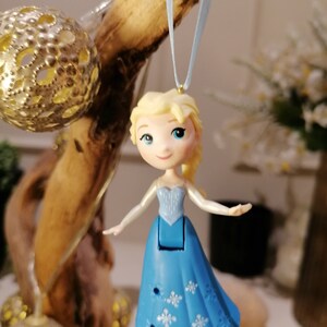 May include: A blue and white plastic ornament of Elsa from Frozen, with a light blue ribbon for hanging. The ornament is a small, detailed figure of Elsa in her signature ice blue dress with a snowflake pattern.