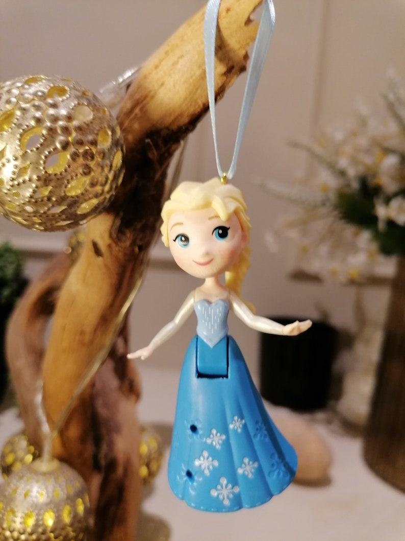 May include: A blue and white plastic ornament of Elsa from Frozen, with a light blue ribbon for hanging. The ornament is a small, detailed figure of Elsa in her signature ice blue dress with a snowflake pattern.