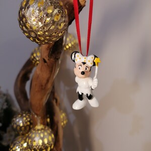 May include: A white ceramic Christmas bauble featuring a cartoon mouse wearing a red bow and holding a star-shaped wand. The bauble is hanging from a red ribbon on a wooden branch.