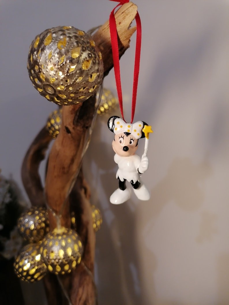 May include: A white ceramic Christmas bauble featuring a cartoon mouse wearing a red bow and holding a star-shaped wand. The bauble is hanging from a red ribbon on a wooden branch.