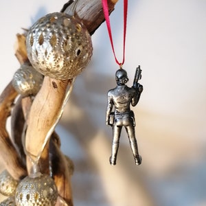 May include: A silver-coloured ornament of a person holding a gun, hanging from a red ribbon on a branch with gold-coloured ornaments.